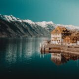 white and brown concrete building beside body of water near snow capped rocky mountain under clear blue sky