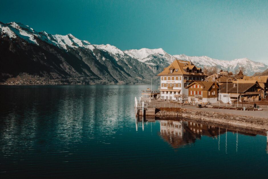white and brown concrete building beside body of water near snow capped rocky mountain under clear blue sky